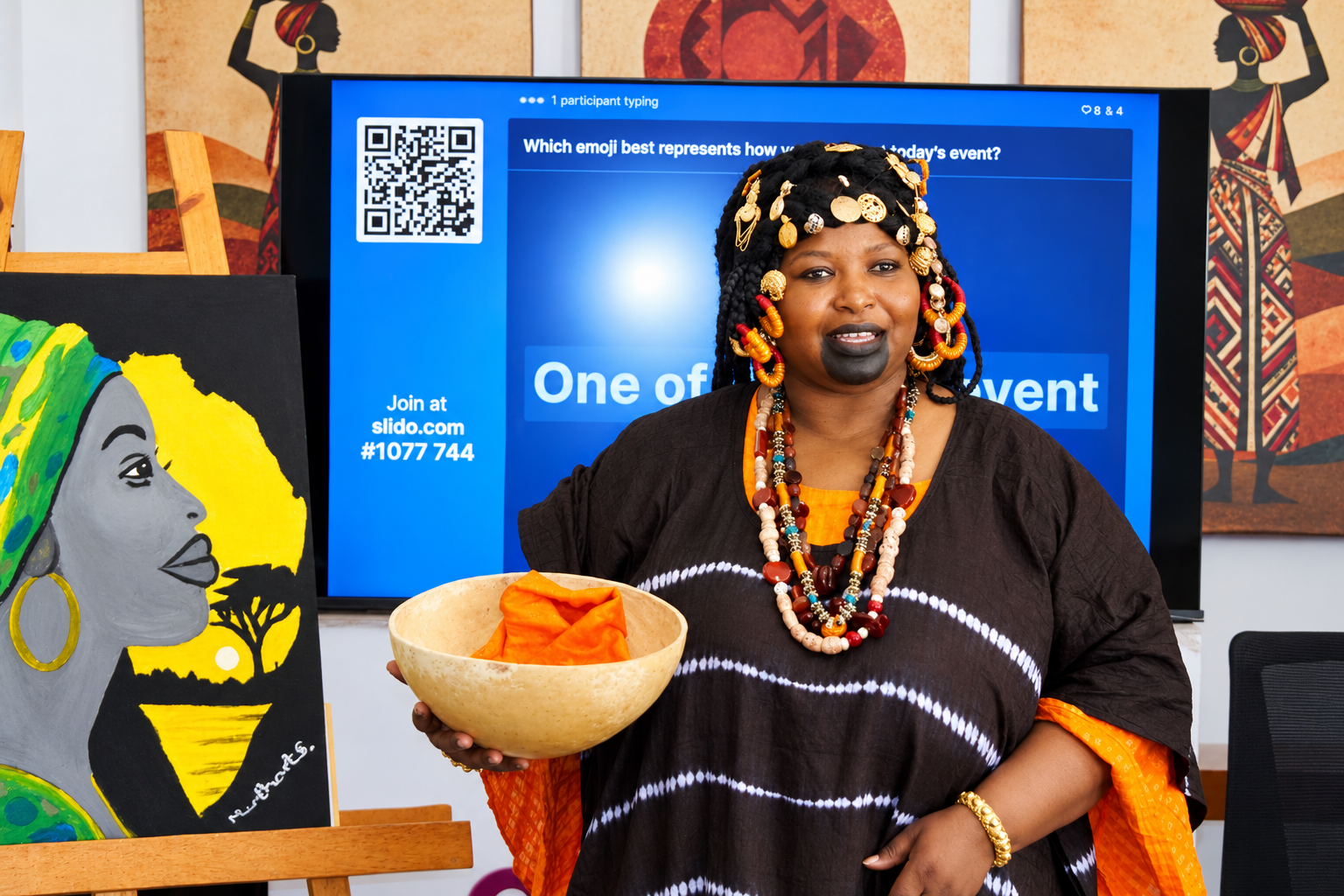 A cultural presenter wearing beaded jewellery and holding a calabash beside African artwork